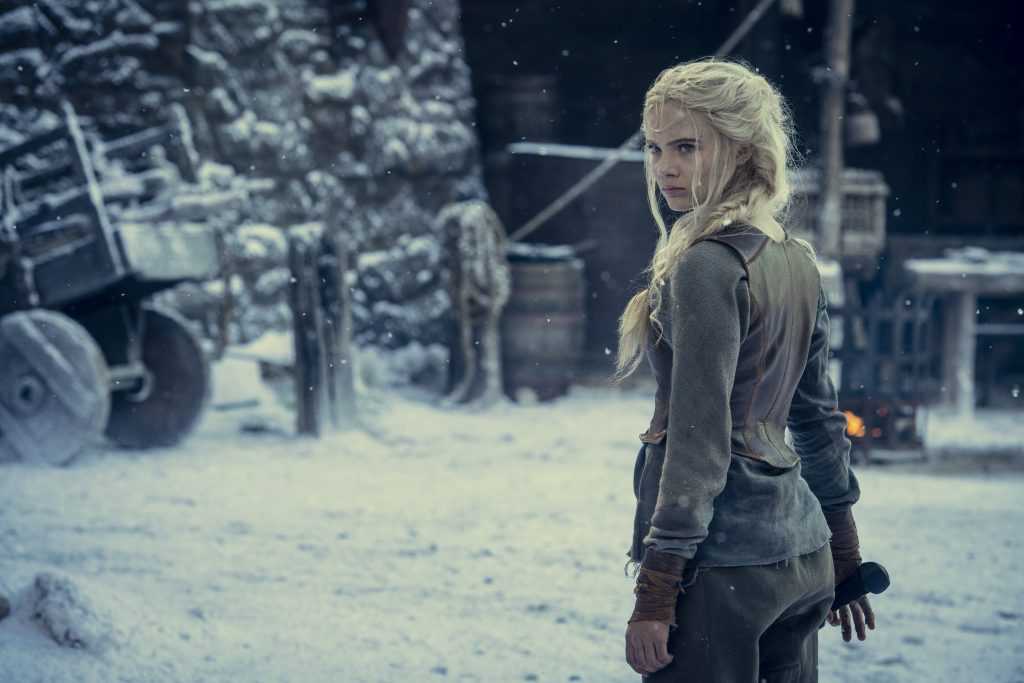 Freya Allan as Princess Ciri from The Witcher. Standing in a snowy set, the character has her back to the camera and is grabbing a sword. It looks like she's training
